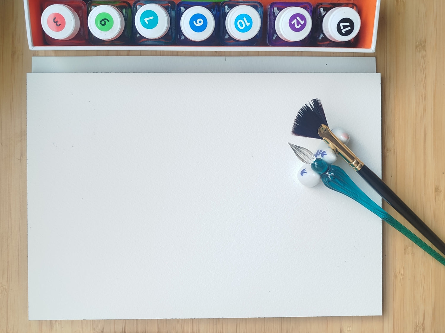 L'image représente une feuille blanche avec un pinceau et une plume pour encre de Chine, ainsi qu'une rangée de flacon d'encres de couleur. C'est une photographie en provenance de l'atelier de l'artiste peintre Maeva J.