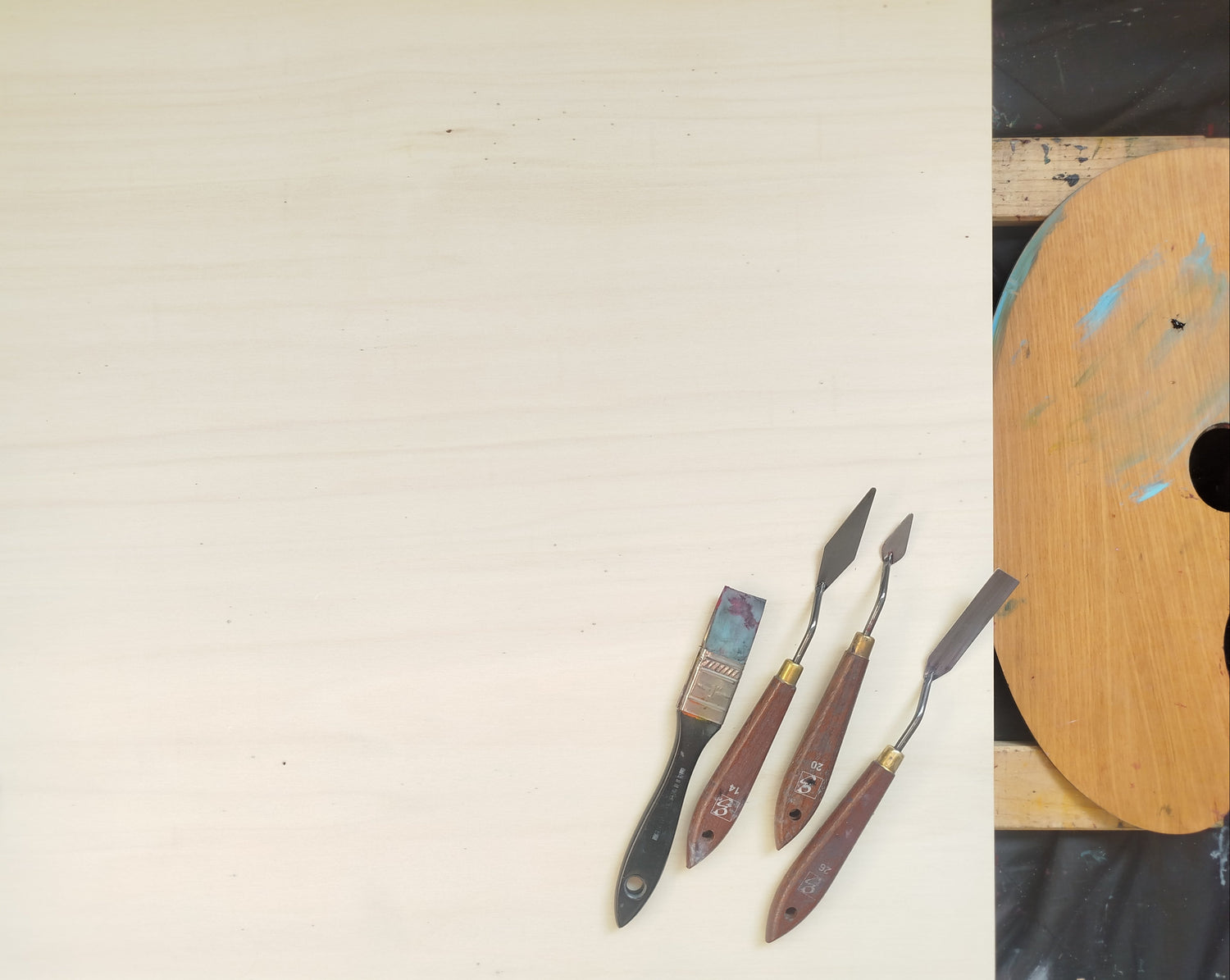 L'image représente un panneau de bois qui est le support de peinture de l'artiste. Sont également visible des couteaux à peindre, un pinceau en silicone, et une palette. C'est une photographie en provenance de l'atelier de l'artiste peintre Maeva J.